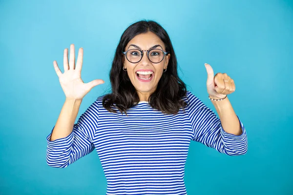 Gözlüklü güzel bir kadın mavi arka planda duruyor. Altıncı parmaklarıyla işaret ediyor ve gülümsüyor. Kendinden emin ve mutlu..