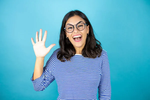 Gözlüklü güzel bir kadın mavi arka planda duruyor. Beş numaralı parmaklarıyla işaret ediyor ve gülümsüyor. Kendinden emin ve mutlu..