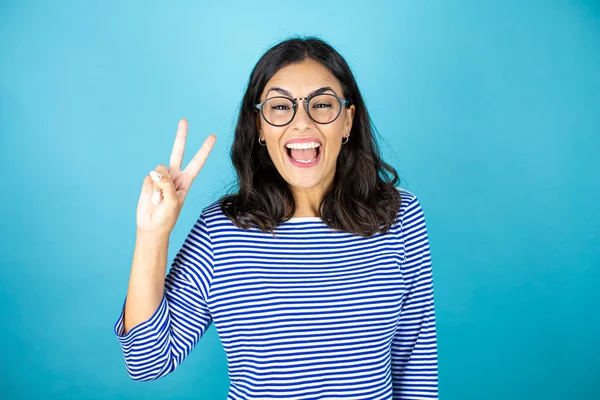 Gözlüklü güzel bir kadın mavi arka planda duruyor. İki numaralı parmaklarıyla işaret ediyor ve gülümsüyor. Kendinden emin ve mutlu.