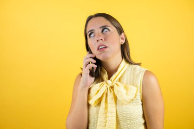 Soyutlanmış sarı arka planda genç ve güzel bir kadın telefonda şaşkın bir ifadeyle konuşuyor.