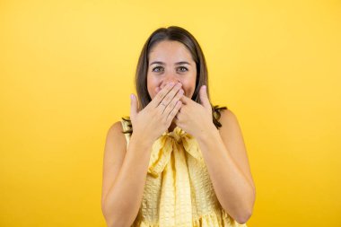 İzole edilmiş sarı arka planda genç ve güzel bir kadın yanlışlıkla ağızlarını kapatarak şok geçirdi. Gizli konsept.