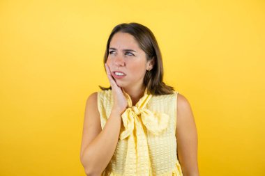 İzole edilmiş sarı arka planda genç bir kadın diş ağrısı ya da diş hastalığından dolayı acı dolu bir ifadeyle dudaklarına dokunuyor.