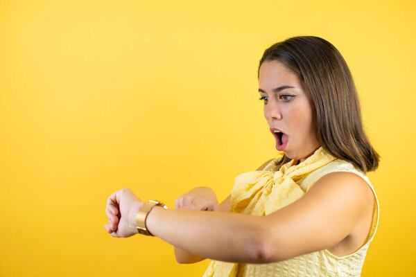 Young beautiful woman over isolated yellow background surprised and pointing his watch because it's late