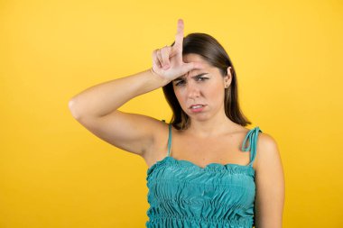 Genç ve güzel bir kadın, izole edilmiş sarı arka planda parmakları alnında insanlarla alay ediyor. Ezik hareketler yapıyor, alay ediyor, hakaret ediyor..
