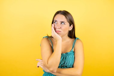 Soyutlanmış sarı arka planı olan genç ve güzel bir kadın çapraz kollardan sıkılmış ve yorgun göründüğünü düşünüyor.