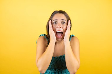Sarı arka planda yaşayan genç ve güzel bir kadın çığlık atıyor ve elleri ağzında şaşırıyor.