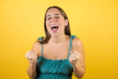 Genç ve güzel bir kadın, izole edilmiş sarı arka planda çok mutlu ve heyecanlı. Kollarını kaldırıp, gülümseyerek ve başarı için çığlık atarak kazanan bir jest yapıyor..