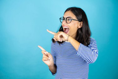 Gözlüklü güzel bir kadın mavi arka planda duruyor. Şaşırmış ve eli ve parmağı yanda.