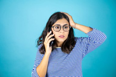 Gözlüklü güzel bir kadın mavi arka planda dikilip telefonda endişeli bir ifadeyle konuşuyor.
