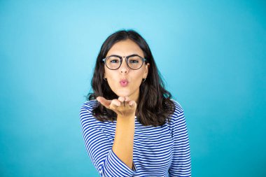 Gözlük takan güzel bir kadın mavi arka planda dikilip kameraya bakıp havada öpüşürken güzel ve seksi görünüyor. Aşk ifadesi.