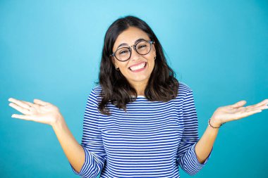 Güzel bir kadın mavi arka plan üzerinde duran gözlük takıyor. Kolları ve elleri havada, kafası karışmış bir ifade.