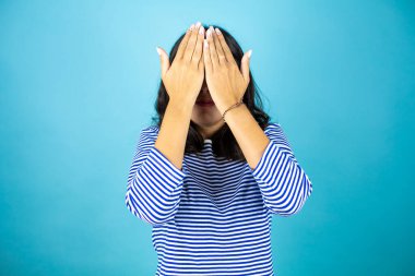 Gözlüklü güzel bir kadın mavi arka planda dikiliyor. Gözleri kollarıyla kaplıyor. Neşeli ve komik gülümsüyor. Kör kavram.