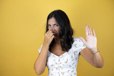 Beautiful woman over yellow background smelling something stinky and disgusting, intolerable smell, holding breath with fingers on nose clipart