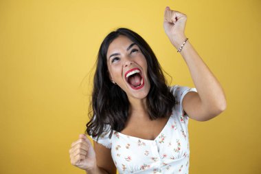 Sarı arka planda güzel bir kadın. Çok mutlu ve heyecanlı. Kollarını kaldırmış, gülümseyen ve başarı için çığlık atan kazanan bir jest yapıyor..
