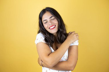 Sarı arka planda güzel bir kadın kendine sarılıyor mutlu ve pozitif, kendinden emin gülümsüyor.