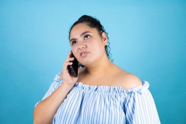 Kıvırcık saçlı, soyutlanmış mavi arka planı olan genç ve güzel bir kadın telefonda kafası karışık bir ifadeyle konuşuyor.