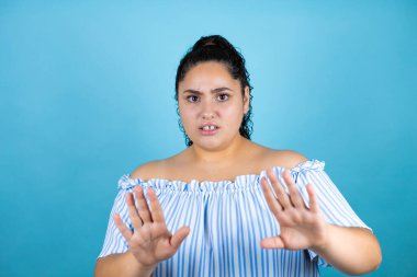 Kıvırcık saçlı, soyutlanmış mavi arka planı olan, korku ifadesinden korkan ve dehşete düşmüş genç bir kadın. El kol hareketi yapmayı kes, şok içinde bağır. Panik konsepti.