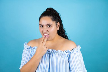 Kıvırcık saçlı, soyutlanmış mavi arka planda eli ağzında genç ve güzel bir kadın.