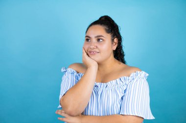 Kıvırcık saçlı, soyutlanmış mavi arka planı olan genç ve güzel bir kadın kollarını kavuşturmaktan sıkılmış ve yorgun görünüyor.