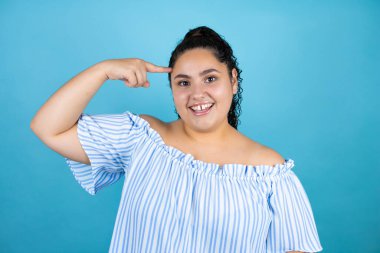 Kıvırcık saçlı, soyutlanmış mavi arka planda genç bir kadın. Gülümsüyor ve parmakları kafasında bir fikri olduğunu düşünüyor..