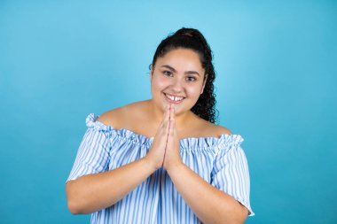 Kıvırcık saçlı, soyutlanmış mavi arkaplan üzerinde genç ve güzel bir kadın elleri bağlı yalvarıyor ve dua ediyor. Yüzünde umut ifadesi var. Çok duygusal ve endişeli.