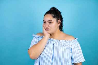 Kıvırcık saçlı, yalıtılmış mavi arka planda genç bir kadın diş ağrısı ya da diş ağrısı yüzünden elleriyle dudaklarına dokunuyor.