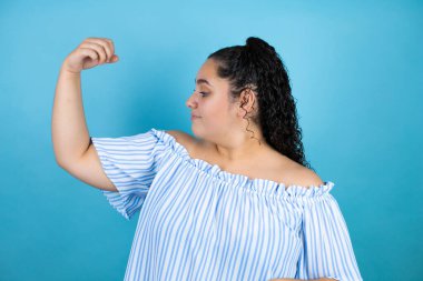 Mavi arka planda kıvırcık saçlı güzel bir kadın gururla gülümseyen kol kaslarını gösteriyor.