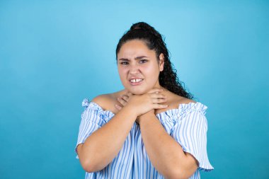 Kıvırcık saçlı, soyutlanmış mavi arka planda genç bir kadın ağrılı boynuna dokunuyor, grip, mantar ve enfeksiyon yüzünden boğazı ağrıyor.