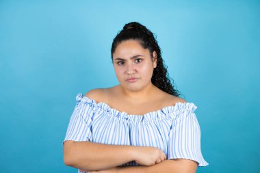 Genç, kıvırcık saçlı, soyutlanmış mavi arka planda kuşkucu ve gergin, yüzünde çapraz kollu tasvip etmeyen bir ifade olan güzel bir kadın.