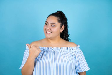 Kıvırcık saçlı, soyutlanmış mavi arka planı olan genç ve güzel bir kadın baş parmağını yan tarafa kaldırıp gülümsüyor.