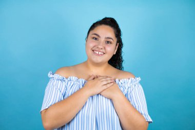 Genç, kıvırcık saçlı, soyutlanmış mavi arka planda elleri göğsünde ve yüzünde minnettarlıkla gülümseyen güzel bir kadın..