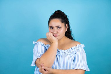 Kıvırcık saçlı, soyutlanmış mavi arka planı olan genç ve güzel bir kadın kollarını kavuşturmaktan sıkılmış ve yorgun görünüyor.
