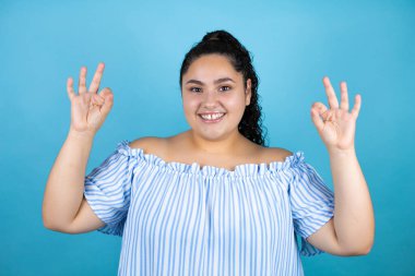 Kıvırcık saçlı, soyutlanmış mavi arka planda genç bir kadın tamam işareti yapıyor parmaklarıyla ve gülümsüyor, mükemmel bir sembol