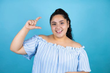 Kıvırcık saçlı, soyutlanmış mavi arka planda gülümseyen ve kendine güvenen, el hareketleriyle küçük boy işareti yapan, parmakları ve kamerası olan güzel bir kadın. Ölçü kavramı.