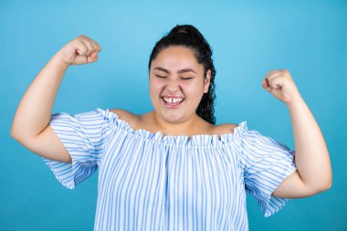 Kıvırcık saçlı, soyutlanmış mavi arka planda çok mutlu ve heyecanlı genç bir kadın kollarını kaldırmış, gülümseyen ve başarı için çığlık atan, kazanan bir jest yapıyor..