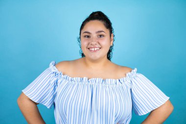 Kıvırcık saçlı, soyutlanmış mavi arka planda kolları belinde gülümseyen güzel bir kadın.