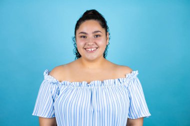 Kıvırcık saçlı, soyutlanmış mavi arka planda gülümseyen ve kameraya bakan güzel bir kadın.