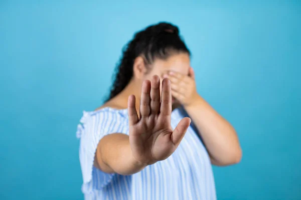 Young beautiful woman with curly hair over isolated blue background covering eyes with hands and doing stop gesture with sad and fear expression. Embarrassed and negative concept.