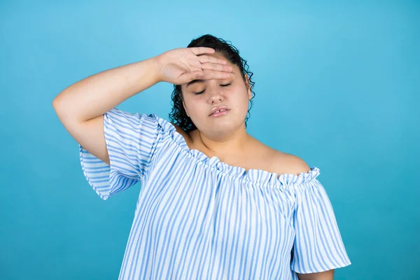 Kıvırcık saçlı, soyutlanmış mavi arka planda genç bir kadın. Hastalık ve ateş için alnına dokunuyor. Grip ve soğuk algınlığı.