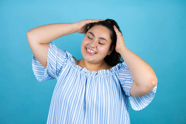 Kıvırcık saçlı genç bir kadın izole edilmiş mavi arka planda rahatlıyor ve esniyor, kolları ve elleri başının arkasında mutlu bir şekilde gülümsüyor.