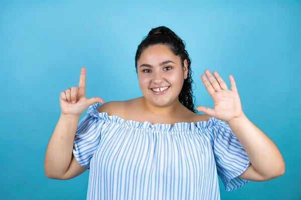 Kıvırcık saçlı, soyutlanmış mavi arka planı olan genç ve güzel bir kadın yedi numaralı parmaklarıyla işaret ederken kendinden emin ve mutlu bir şekilde gülümsüyor.