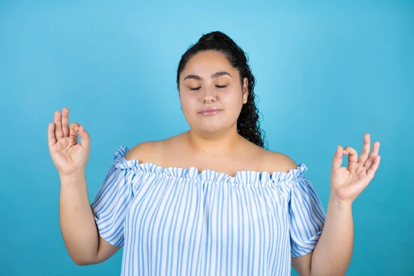 Kıvırcık saçlı genç bir kadın izole edilmiş mavi arka planda rahatla ve gözleri kapalı gülümse. Parmaklarıyla meditasyon hareketi yapıyor.