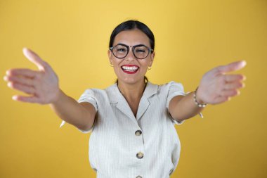 Sarı arka planda güzel bir kadın kameraya bakıyor. Sarılmak için kollarını açmış gülümsüyor.