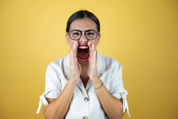 Beautiful woman over yellow background shouting and screaming loud to side with hands on mouth