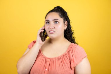 Sarı arka plan üzerinde kıvırcık saçlı genç ve güzel bir kadın telefonda şaşkın bir ifadeyle konuşuyor.