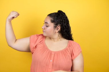 Sarı arka planda kıvırcık saçlı güzel bir kadın gururla gülümseyen kol kaslarını gösteriyor.