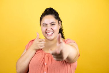 Sarı arka planda kıvırcık saçlı, genç ve güzel bir kadın seni ve kamerayı işaret ediyor.