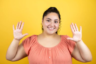 Sarı arka planda kıvırcık saçları olan genç ve güzel bir kadın on numarayı işaret ederken kendinden emin ve mutlu bir şekilde gülümsüyor.