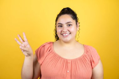 Sarı arka planda kıvırcık saçlı genç bir kadın iki numaralı parmaklarıyla gülümseyerek kendinden emin ve mutlu olduğunu gösteriyor.