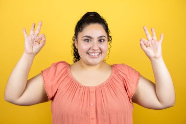Sarı arka planda kıvırcık saçlı güzel bir kadın. Tamam işareti yapıyor. Parmaklarıyla ve gülümsüyor.